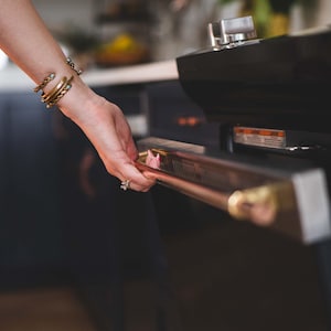 May include: A close-up of a black oven with a gold-toned handle. The oven door is being opened. The handle is a long, horizontal bar. The oven is in a kitchen setting. A person's arm is visible, adorned with gold bracelets.