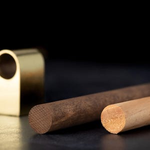 May include: A close-up shot features a gold-colored, arched metal piece with a circular opening, alongside two wooden dowels. One dowel is a dark brown color, and the other is a light tan color. The background is black.