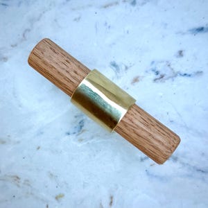 May include: A wooden cabinet pull with a brass band. The pull is made of light brown wood and has a cylindrical shape. The brass band is a shiny gold color and wraps around the center of the wooden pull.