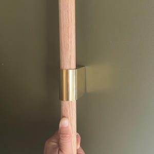 May include: A cylindrical light-toned wooden handle secured by a polished gold-colored metal bracket. The hardware features a smooth, vertical design that connects the wooden rod to a flat surface, showcasing a clean and functional aesthetic.