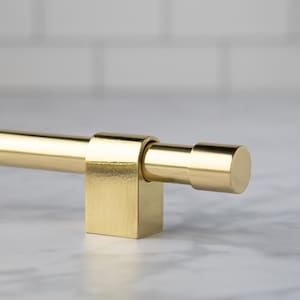 May include: A close-up of a gold-colored cabinet pull. The pull has a cylindrical bar and a square base with a cylindrical end. The hardware is made of metal and has a brushed finish. The background is a white tile.