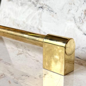 May include: A polished brass towel bar featuring a cylindrical horizontal rod connected to a rectangular mounting bracket with a circular end cap. The metal surface has a reflective gold-toned finish with subtle variations in the brass texture.
