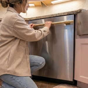 May include: A stainless steel dishwasher featuring a long, horizontal, cylindrical brass handle. The appliance has a metallic finish and is installed between light pink kitchen cabinets with a granite countertop above it.