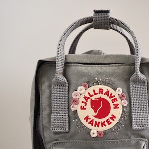 May include: A gray Fjällräven Kånken backpack with a white embroidered floral design and the Fjällräven logo in red. The backpack has adjustable straps and a top handle.