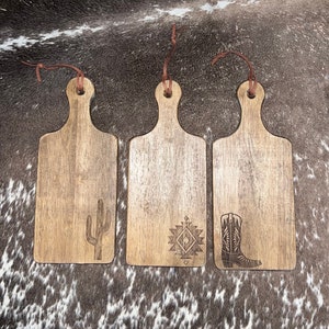 May include: Three wooden cutting boards with leather straps. The boards are decorated with a cactus, a geometric pattern, and a cowboy boot.