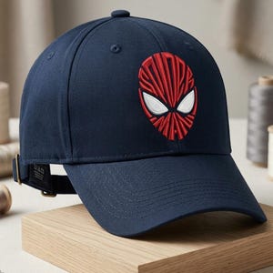 Könnte beinhalten: Eine marineblaue Baseballkappe mit einer gestickten rot-weißen Grafik einer Maske auf der Vorderseite. Das Design integriert den Text "SPIDER-MAN" in die rote Maskenform. Die Kappe verfügt über einen gebogenen Schirm und einen verstellbaren Riemen.