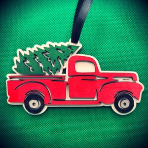 May include: Red vintage pickup truck with a green Christmas tree in the bed. The truck is a wooden ornament with a black ribbon for hanging.