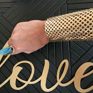 May include: A gold metal cuff bracelet with a circular pattern. The bracelet is on a person's arm. The person is wearing a ring with a blue gemstone.