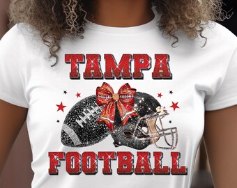 Puede incluir: Camiseta blanca con las palabras "TAMPA FOOTBALL" en letras rojas brillantes. La camiseta presenta un diseño de fútbol americano y casco, adornado con un lazo rojo y estrellas, creando un tema deportivo.