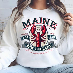 May include: White sweatshirt with a graphic design featuring a red lobster, the words "Maine", "Coastal Est 1820", "New England", and "Fresh Lobster", and the text "Maine" in a circle.