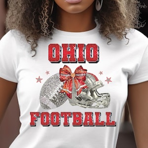 May include: White t-shirt with a glitter design. The word "OHIO" is above a football, helmet, and a red bow. The word "FOOTBALL" is below in red glitter. Small red stars are scattered around the design.
