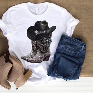 May include: White t-shirt with a black cowboy hat and boots with rhinestones. The boots have a black and white floral design. The shirt is laying on a brown surface with a pair of brown ankle boots and blue jeans.