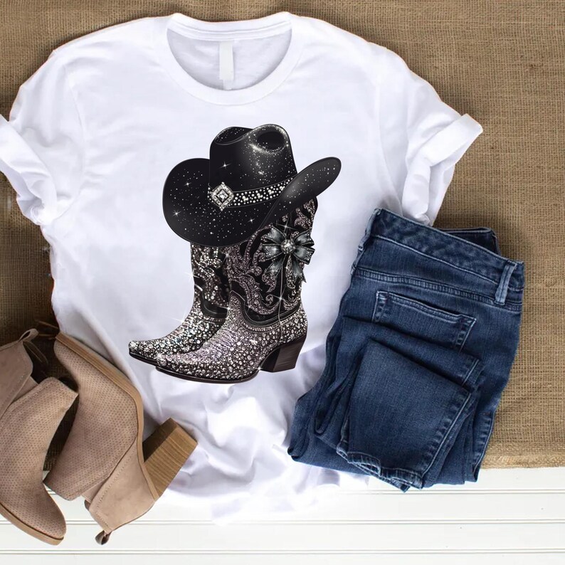 May include: White t-shirt with a black cowboy hat and boots with rhinestones. The boots have a black and white floral design. The shirt is laying on a brown surface with a pair of brown ankle boots and blue jeans.