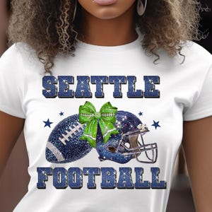 May include: White t-shirt with the words "SEATTLE FOOTBALL" in blue glitter lettering. The shirt features a football, a helmet, and a green bow, all decorated with blue glitter and stars.