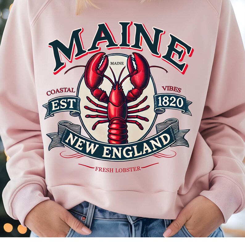 May include: A pink sweatshirt with a graphic design featuring a lobster, the words "Maine", "Coastal Est 1820 Vibes", "New England", and "Fresh Lobster".