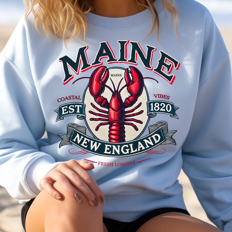 May include: Light blue sweatshirt with a graphic design featuring a red lobster, the text "Maine", and the words "Coastal Est 1820 Vibes New England Fresh Lobster".