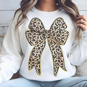 May include: White sweatshirt with a large leopard print bow design. The bow is outlined in gold glitter.