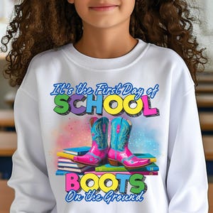 May include: White sweatshirt with the text "It's the First Day of SCHOOL BOOTS On the Ground." The graphic features a stack of books and a pair of pink and blue cowboy boots.