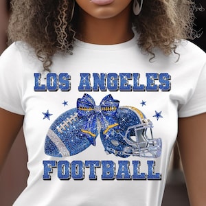 May include: White t-shirt with a blue and silver design featuring a football, helmet, and bow. The text "LOS ANGELES FOOTBALL" is displayed in blue. The design includes star accents.