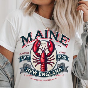 May include: White t-shirt with a graphic design featuring a red lobster, the words "Maine", "Coastal Est 1820", "New England", and "Fresh Lobster".