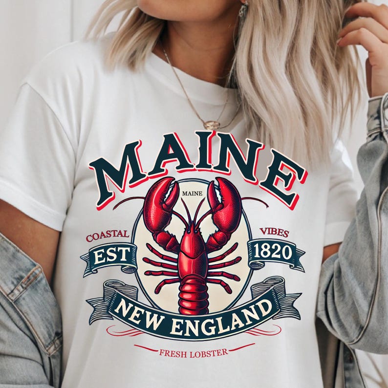May include: White t-shirt with a graphic design featuring a red lobster, the words "Maine", "Coastal Est 1820", "New England", and "Fresh Lobster".