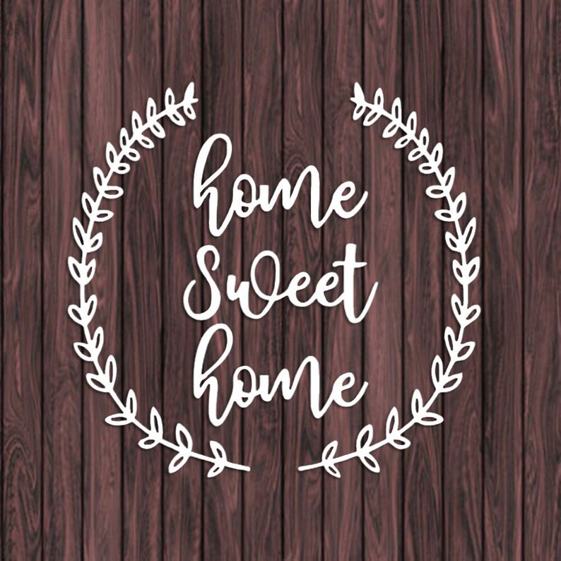 May include: White text on a brown wood background reads "home sweet home" in a cursive font. The text is surrounded by a white wreath of leaves.