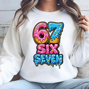 May include: White sweatshirt with a colorful graphic of the numbers "67" designed as donuts with pink frosting and blue icing. The word "SIX" is in pink, and "SEVEN" is in blue, with a dripping icing effect. The sweatshirt is a casual style.