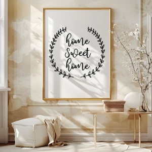 May include: A framed print with a white background and black text that reads "home sweet home" surrounded by a wreath of leaves. The print is hanging on a beige wall.