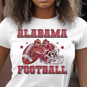 May include: White t-shirt featuring the words "ALABAMA FOOTBALL" in red glitter lettering. The design includes a red glitter football, a matching helmet, and red star accents. The shirt is a classic crew neck style.