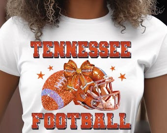 Może przedstawiać: Biała koszulka z napisem "TENNESSEE FOOTBALL" w pomarańczowym brokatowym napisie. Projekt zawiera piłkę nożną, kask i gwiazdy, wszystkie w pomarańczowym brokatowym kolorze. Piłka ma fioletowy pasek i kokardę.
