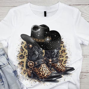 Könnte beinhalten: Weißes T-Shirt mit einem Aufdruck von zwei schwarzen Cowboystiefeln mit Leopardenmuster und einem schwarzen Cowboyhut mit silbernen Sternen.