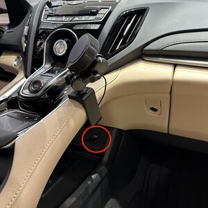 Puede incluir: Primer plano del interior de un coche negro con un soporte para tel&eacute;fono montado en el salpicadero. Un bot&oacute;n negro con un s&iacute;mbolo es visible en la consola central.
