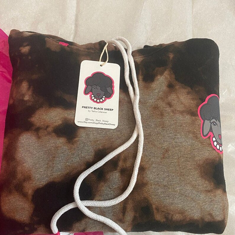 May include: A brown and black tie-dye hoodie with a white drawstring. The hoodie features a black sheep graphic with a pink bow and white pearls. The tag on the hoodie says "Pretty Black Sheep by Tasha Celener" and includes the social media handle "@Pretty_Black_Sheep" and website "www.etsy.com/shop/PrettyBlackSheep".