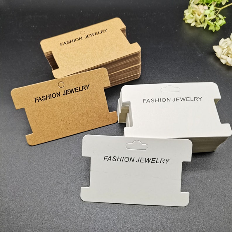 Puede incluir: Una pila de tarjetas de exhibici&oacute;n de aretes de cart&oacute;n marr&oacute;n y blanco con el texto "FASHION JEWELRY" impreso en cada tarjeta.