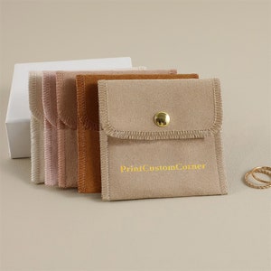 May include: Five small, square, beige suede pouches with a gold button closure. The pouch on the right has the text "PrintCustomCorner" printed in gold on the front.
