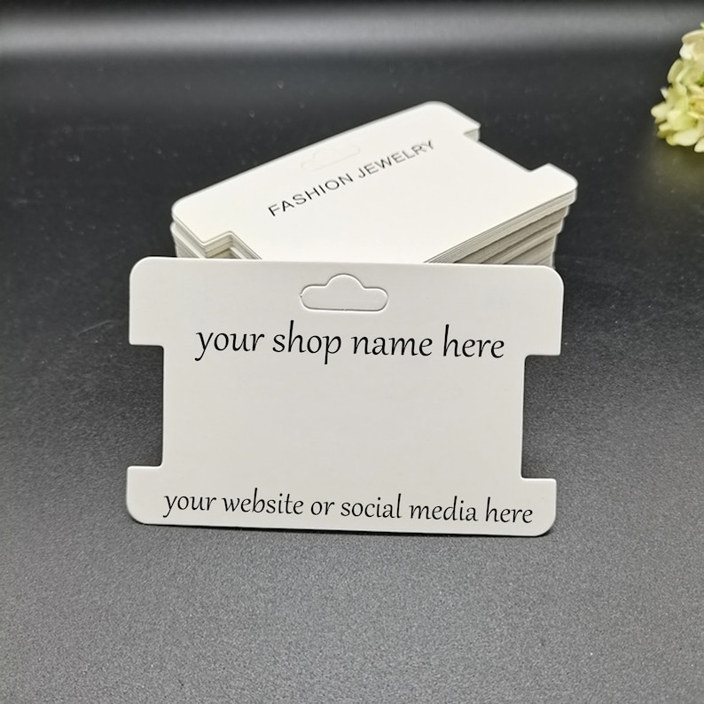 Puede incluir: Etiquetas colgantes de cartulina blanca con una ranura para un collar o una pulsera. Las etiquetas tienen el texto "FASHION JEWELRY" en la parte superior y el texto "your shop name here" y "your website or social media here" en la parte inferior.