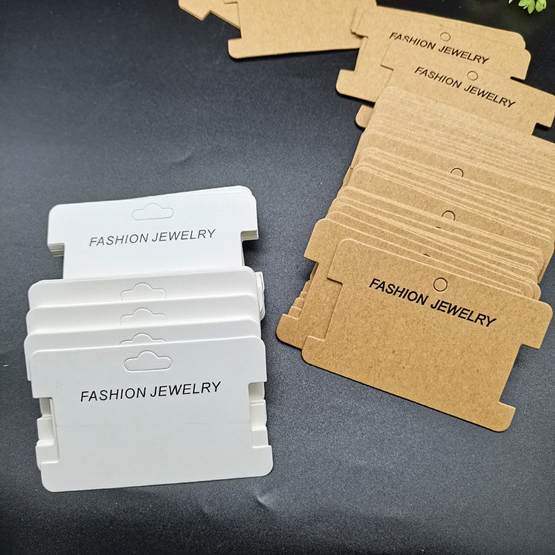 Puede incluir: Una pila de etiquetas rectangulares blancas y marrones con el texto "FASHION JEWELRY" impreso en ellas. Las etiquetas tienen un agujero en la parte superior para colgar.