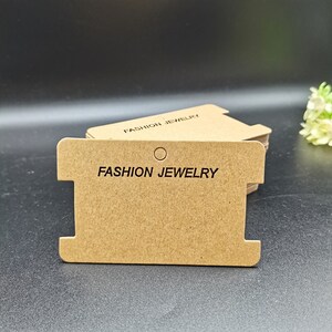 Puede incluir: Una pila de etiquetas de joyer&iacute;a de cart&oacute;n marr&oacute;n con un agujero en la parte superior y una ranura a cada lado. Las etiquetas est&aacute;n impresas con el texto "FASHION JEWELRY".