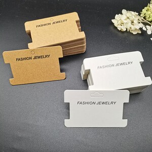Puede incluir: Dos pilas de etiquetas de joyer&iacute;a en blanco, una marr&oacute;n y otra blanca. Las etiquetas tienen un agujero en la parte superior y una ranura en la parte inferior. El texto "FASHION JEWELRY" est&aacute; impreso en cada etiqueta.