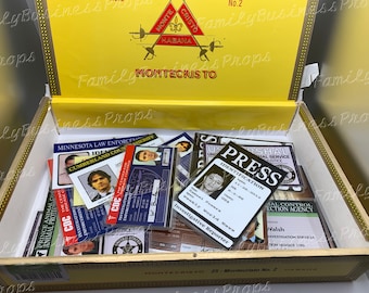 May include: A wooden cigar box with a yellow lid and gold trim. The lid has the words "Montecristo No. 2" and "Habana" printed on it. The box is open and filled with various fake identification cards, including a "Press" card with a photo of a man and the words "Investigative Reporter".