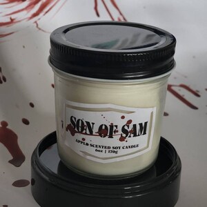 Può includere: Una candela in un barattolo di vetro con un coperchio nero e un'etichetta bianca che dice "Son of Sam Apple Scented Soy Candle 6oz | 170g".