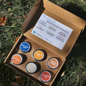 May include: A cardboard box with a selection of six small candle tins. The tins are arranged in two rows of three. The candle scents are: Fall Night Long, Bonfire, Pumpkin Pie, Maple Pecan, Cinnamon Spice, and Hot Apple Pie. The box has a label that says "Fall Sampler Set".