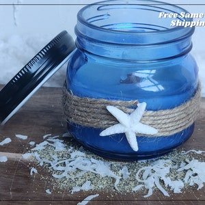 Puede incluir: Un tarro de vidrio azul con una estrella de mar blanca y cuerda envuelta alrededor. El tarro está sentado sobre una superficie de madera con coco rallado esparcido alrededor.
