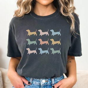 May include: A dark grey t-shirt featuring a graphic of several pastel-coloured dachshund silhouettes. The text "PEPPER" is in the top left corner of the image.