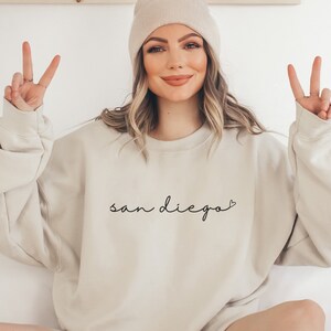 May include: A white sweatshirt with the text "san diego" in black script with a small heart after the "o". The model is wearing the sweatshirt and has a peace sign with each hand.
