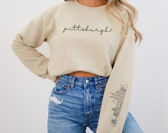 Peut inclure: Un sweat-shirt beige clair avec le mot "pittsburgh" écrit en cursive sur le devant. Le sweat-shirt a un graphique de skyline sur la manche gauche. Le mannequin porte un jean bleu.