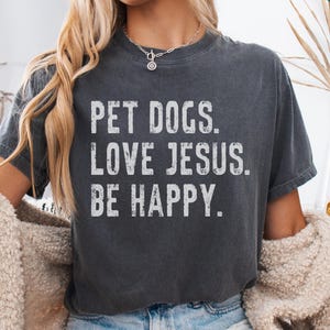 May include: A dark gray t-shirt with the text "Pet Dogs. Love Jesus. Be Happy." printed in white. The shirt is being modeled by a person. The text "Medium Pepper" is visible at the bottom of the image.