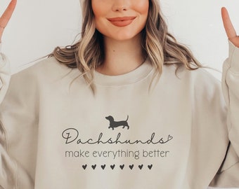 May include: A beige sweatshirt with the text "Dachshunds make everything better" and a small silhouette of a dachshund. The sweatshirt has a crew neck and long sleeves. The text is in black and the silhouette is in gray. The sweatshirt is on a model wearing a white beanie.