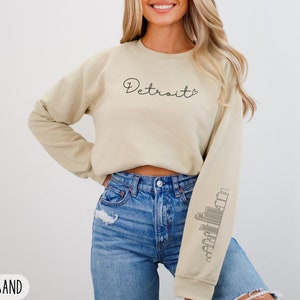 May include: A beige sweatshirt with the word "Detroit" written in black cursive on the front. The sweatshirt has a skyline graphic on the left sleeve. The model is wearing blue jeans.