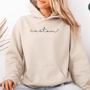 May include: A medium sand-colored hoodie with a front pocket and a drawstring hood. The word "custom" is written in cursive across the chest. The hoodie is worn with blue jeans.
